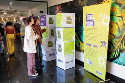 Pessoas a interagir com os elementos informativos da campanha "Giro da Verdade", composta por estruturas em cubo e um banner informativo no espaço do MPF.