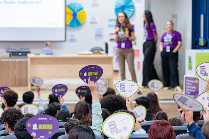 Público num auditório levanta placas ovais em forma de leque, com algumas a exibir a frase "Xô, mentira!", durante uma palestra ministrada por facilitadoras vestidas com camisolas roxas no palco