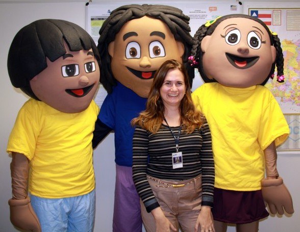 Imagem de uma mulher sorridente ao centro, posando de pé com três mascotes infantis ao seu redor e atrás dela.