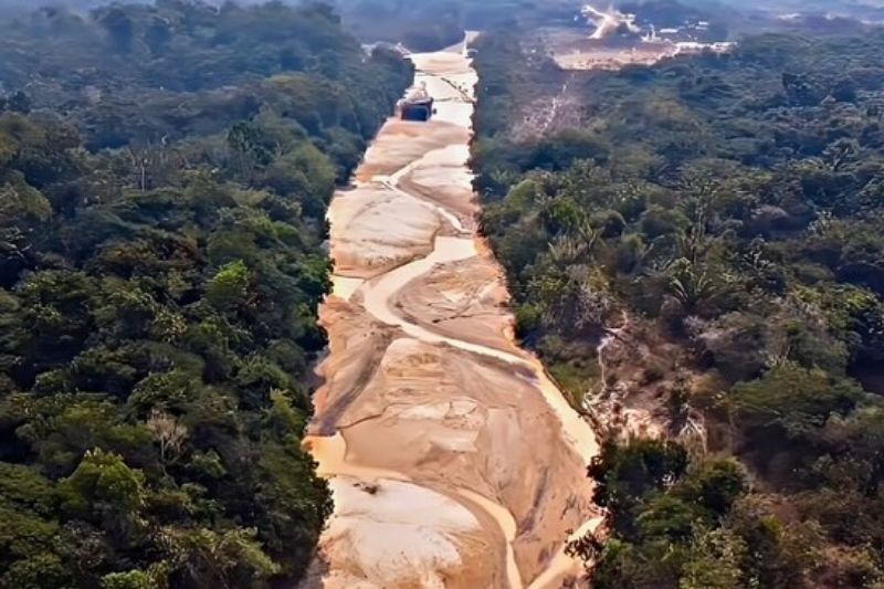 Vista aérea de um rio seco com leito de areia clara e finos filetes de água, cercado por densa floresta tropical verde.