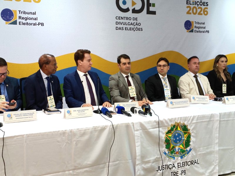 Fotografia de uma coletiva de imprensa em ambiente institucional. Sete pessoas estão sentadas lado a lado atrás de uma mesa longa coberta por toalha branca. Sobre a mesa há microfones, crachás e placas de identificação. Ao fundo, um painel branco exibe os logotipos do Tribunal Regional Eleitoral da Paraíba, do Centro de Divulgação das Eleições e a inscrição Eleições 2026.