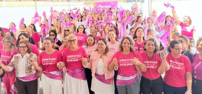 Em um espaço amplo e coberto, dezenas de pessoas participam de um evento da campanha Outubro Rosa. Em primeiro plano, uma fileira de participantes está de mãos dadas. A maioria veste camisetas cor-de-rosa com a frase Cuidar de você é o maior ato de amor. Algumas pessoas usam faixas na cintura também em tom rosa.