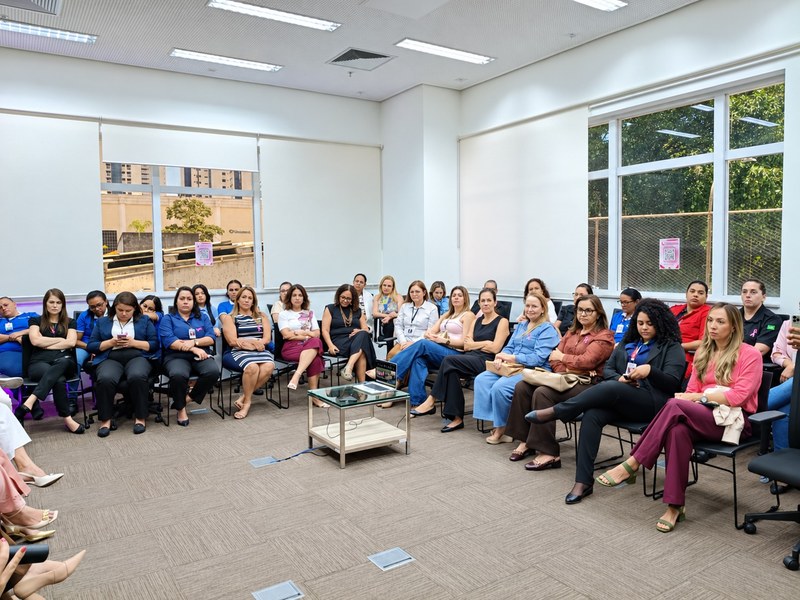Sala de reunião ampla e bem iluminada, com paredes claras, janelas grandes e piso acarpetado. Cerca de trinta pessoas estão sentadas em cadeiras organizadas em semicírculo, voltadas para o centro da sala. No meio, há uma pequena mesa de vidro com um notebook. Algumas pessoas usam roupas com detalhes ou laços cor-de-rosa. Pelas janelas, é possível ver árvores e edifícios ao fundo. O ambiente sugere um encontro ou palestra em grupo.