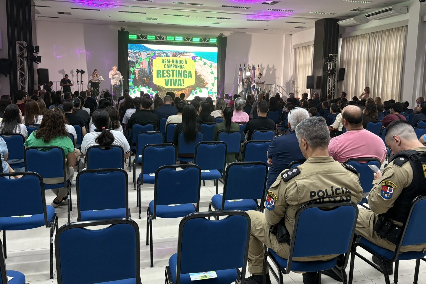 Fotografia em perspectiva frontal de um auditório iluminado durante o lançamento da campanha "Restinga Viva!". Em primeiro plano, no canto inferior direito, estão dois policiais militares fardados sentados de costas, observando o palco. As fileiras de cadeiras azuis estofadas ocupam a maior parte do recinto, onde diversas pessoas assistem ao evento. Ao fundo, um palco iluminado com luzes roxas e verdes exibe um painel de LED central com a imagem de uma praia e o texto em letras garrafais "BEM-VINDO À CAMPANHA RESTINGA VIVA!". No palco, uma pessoa fala ao microfone diante de um púlpito ao lado de um suporte com bandeiras oficiais.