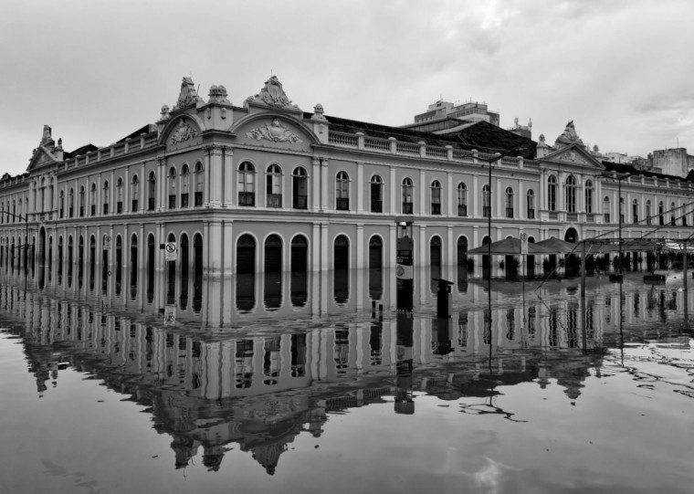 Fotografia em preto e branco de um grande prédio histórico parcialmente alagado por uma enchente. A água cobre a rua até a base dos arcos e portas do pavimento térreo, formando um reflexo lípido da fachada e do céu nublado