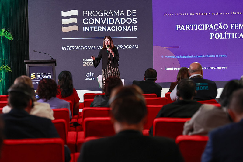 Fotografia de evento em auditório do TSE. Em primeiro plano, vistas de costas, estão várias pessoas sentadas em poltronas vermelhas, acompanhando a apresentação. No centro do palco, em pé, uma mulher de cabelos castanhos na altura dos ombros e óculos fala ao microfone segurando um passador de slides. Ela veste uma blazer preto sobre um vestido longo estampado.
Ao fundo, há painéis em tons de cinza e roxo. No painel da esquerda, lê-se em letras maiúsculas brancas: "PROGRAMA DE CONVIDADOS INTERNACIONAIS" e "INTERNATIONAL VISITORS PROGRAM", acompanhados do logotipo do evento. No painel roxo, à direita, destaca-se o texto: "GRUPO DE TRABALHO VIOLÊNCIA POLÍTICA DE GÊNERO" e o título "PARTICIPAÇÃO FEMININA NA POLÍTICA", além do link institucional do MPF e o nome da palestrante, Raquel Branquinho.