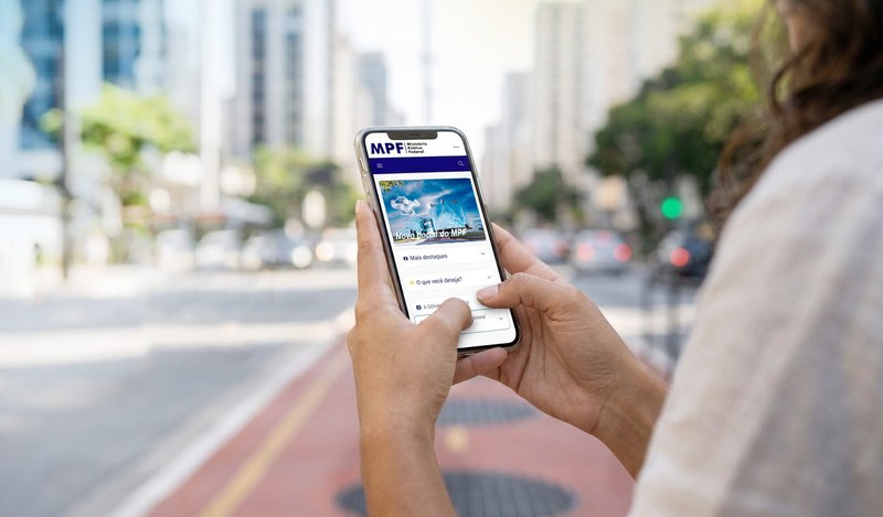 Uma pessoa segura um smartphone e navega no novo portal do Ministério Público Federal (MPF), que exibe um banner sobre tecnologia e acessibilidade para o cidadão. Ao fundo, uma avenida urbana movimentada e prédios altos estão desfocados.