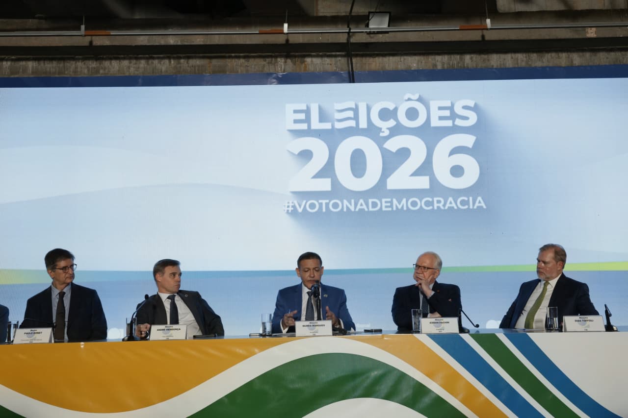 Foto mostra integrantes do TSE e o PGR sentados em uma mesa de coletiva de imprensa. Atrás há um banner onde está escrito Eleições 2026 #votonademocracia